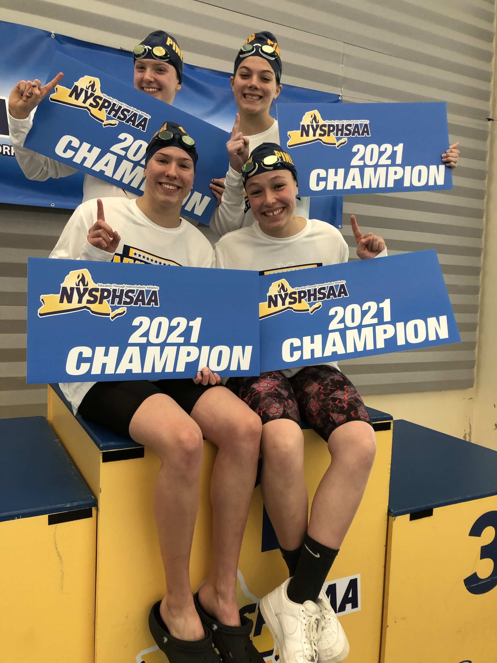 Four young women wearing swim caps, shorts and white tshirts hold banners that say NYSPHSAA 2021 Champion. they are all smiling broadly.