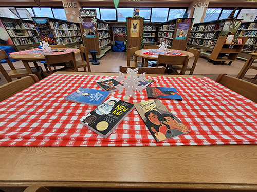 A table in a library with a red and white check table cloth. On it is a cutout of a large snowflake and around that are six  books in a circle.