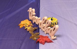 On a light and dark purple color tablecloth is a wooden cutout of the word thankful. In front of it are paper leaves in autumn colors and behind it is a light colored pumpkin.