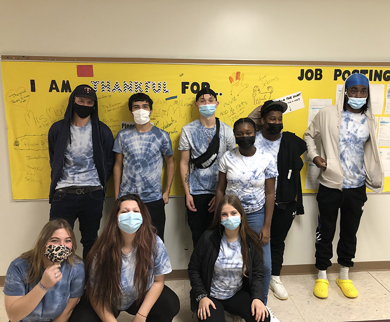 A group of nine high schools students stand in a hallway wearing their blue and white tie dye shirts. All are wearing masks.