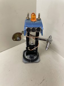 A small sculpture made of different electronics parts. It looks like a tiny robot, with a blue top, three eyes, an orange light on top and holding what looks like a palate and a pen.