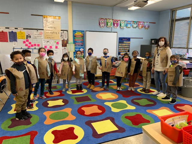 Twelve kindergarten students stand around a classroom, all wearing brown paper vests with a letter of the alphabet on it. There is one adult on the right . All are wearing face masks. They are standing on a multicolored rub with different shapes on it.