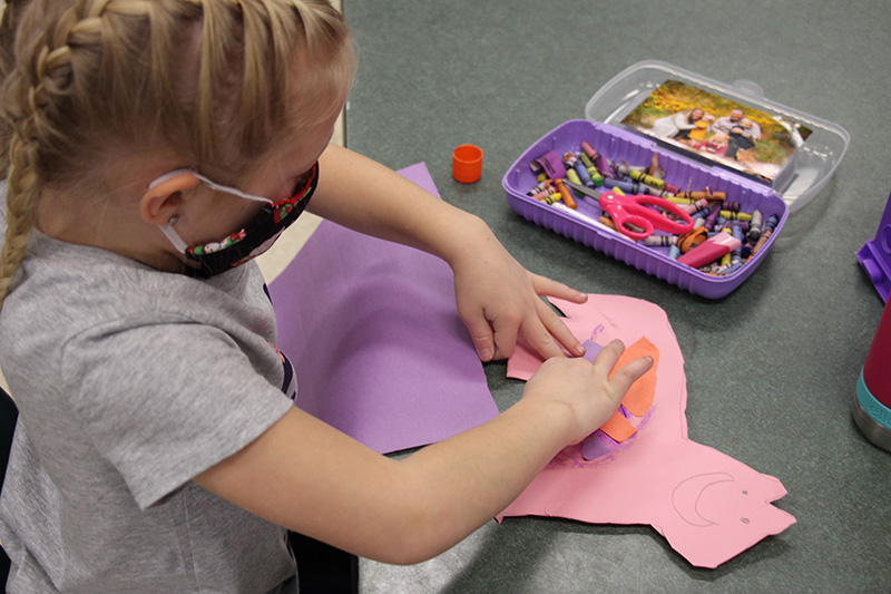 A kindergarten girl with blond hair, pastes pieces of colored paper onto a pink cutout of a horse.