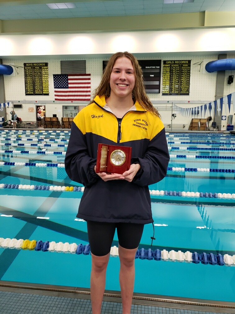 A young woman wearing blue sorts and a warm up jacket in blue and gold smiles and holds an award. Behind her is a large pool with lanes marked off and an American flag.