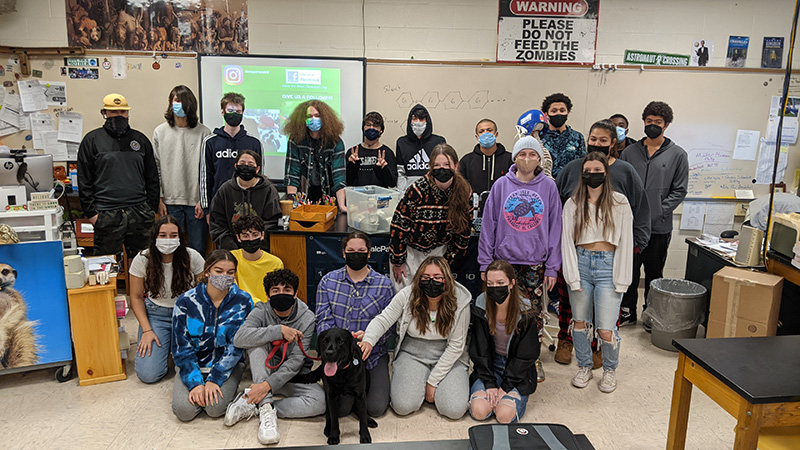 About 20 high school students stand in three rows, classroom in the background. In the center is a black lab. One of the girls in the front is petting the dog. All students have masks on.