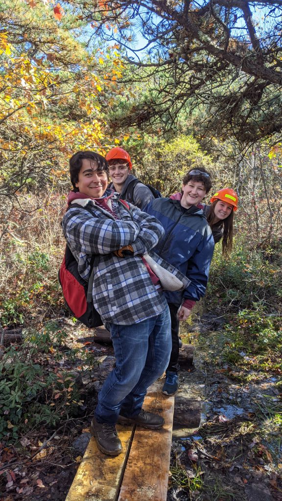 Four students, wearing long sleeve jackets, jeans, backpacks. Two have hardhats on. They are standing on a log, with greenery in the background.