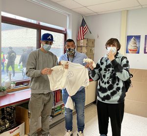 Three high school students hold up their white tshirts that are tied in different places, getting ready to tie dye them. 