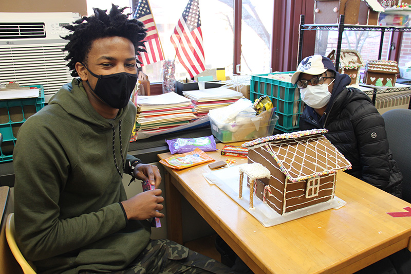 Two young men, high school age, sit at a desk with a gingerbread house on it. The house is very elaborate, with a criss cross pattern on the roof and tiny multi-colored candies outlining it.