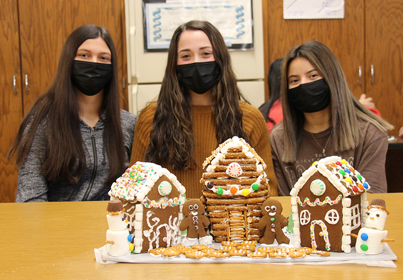 Three young women, all with long dark hair and black masks, sit at a table with three gingerbread houses in front of them. One is larger and has large pretzels all over it as if it were a log cabin. It also has colorful candy around it. The other two are a little smaller but nicely decorated.