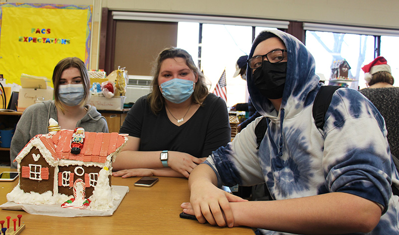 Three high school students sit at a table wearing masks. In front of them is a gingerbread house, nicely decorated with red candies all over the roof.