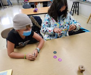 Two elementary students sit at a wooden table watching a dreidel spin. On the table are three different colored coins. The students are wearing masks.