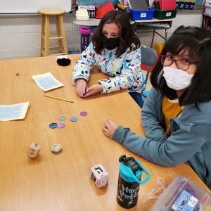 Two girls, both with dark shoulder-length hair and wearing masks sit at a table where there are different colored coins and a dreidel spinning.