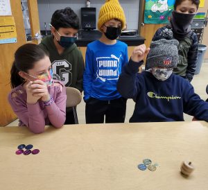 Four elementary students are sitting at a wooden table with different color coins on it. All have masks on and they are watching intently as a dreidel spins. The boy on the right has his arm up, as if he just let go of the dreidel for its spin.