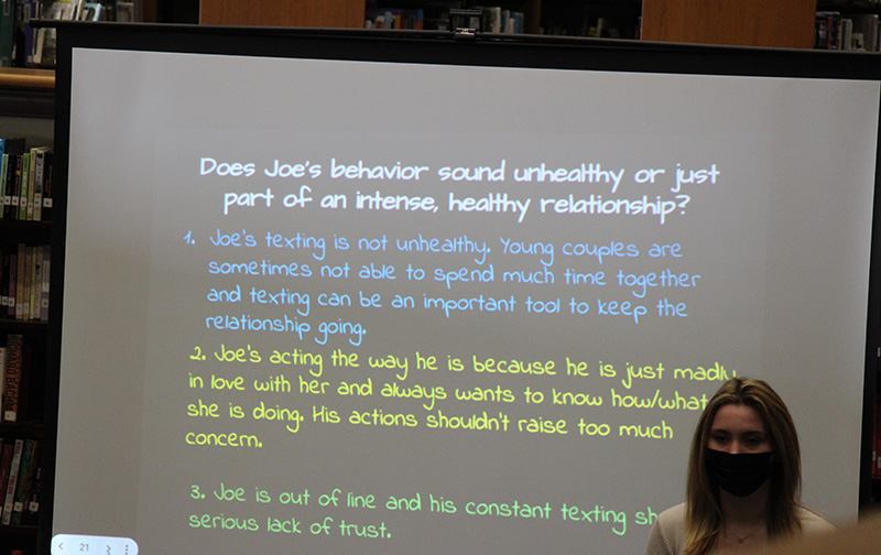 A masked high school student stands next to a screen the multicolored sentences on it.