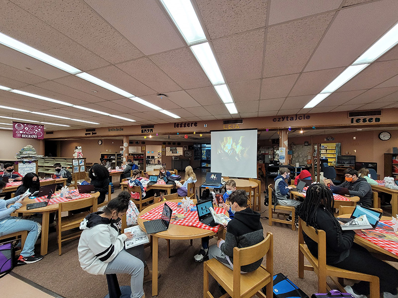 More than a dozen middle school students sitting in a library reading books. They are all masked and are sitting at tables of three, all spaced apart.