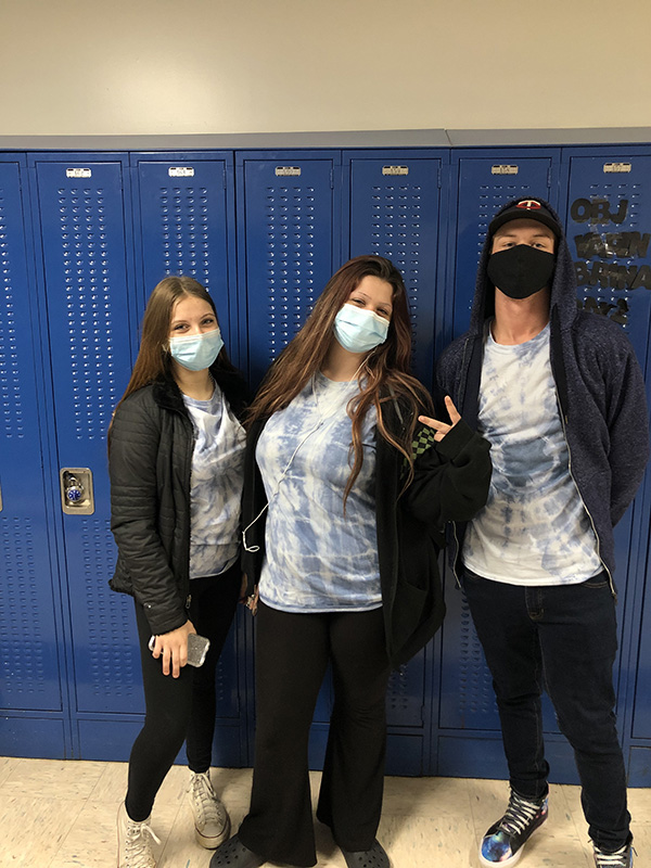 Three high school students stand inf ront of blue lockers. They all have on blue and white tie dye shirts. They are all wearing black jackets and masks. The girl in the center is giving a peace sign.