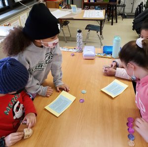 Four elementary students sit around a wood table. They are looking at cards that have instructions on them. There are coins stacked up and a dreidel in one of the students' hands.