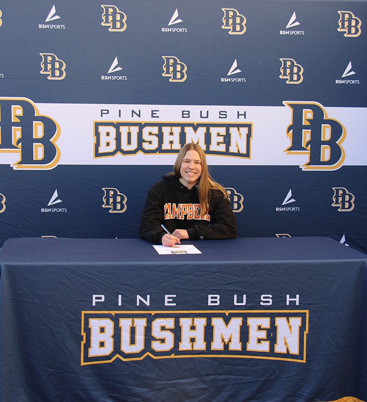 The background is blue and gold with PB Pine Bush Bushmen on the backdrop. Sitting at a table covered in a blue tablecloth is a young woman with long blonde hair. She his holding a pen and signing a piece of paper.