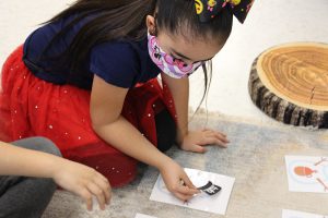 An elementary age girl with long dark hair, wearing a big bow in her hard, kneels on the floor and puts a black top hat on a paper snowman.