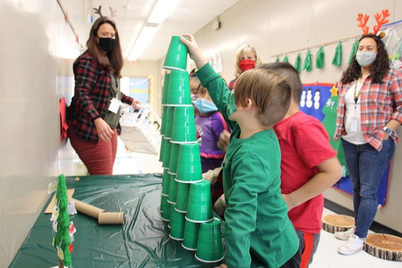 A small group of elementary students stack green plastic cups up high in a pyramid shape. A woman wearing a headband with antlers on it stands and watches. All are wearing masks.