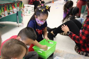 An elementary-age girl with a large bow in her hair, dark hair pulled back, uses tweezers to select a piece of paper from a green bin. She is wearing a purple print mask.