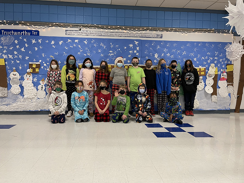 A group of 16 older elementary students in two rows. Behind them is a large bulletin board of a snow scene. All kids are wearing masks.