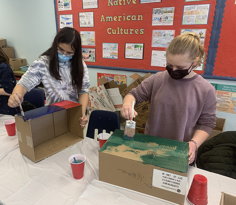 Two middle sch stand and paint cardboard boxes. There are red cups filled with paint. on the table.ool students 