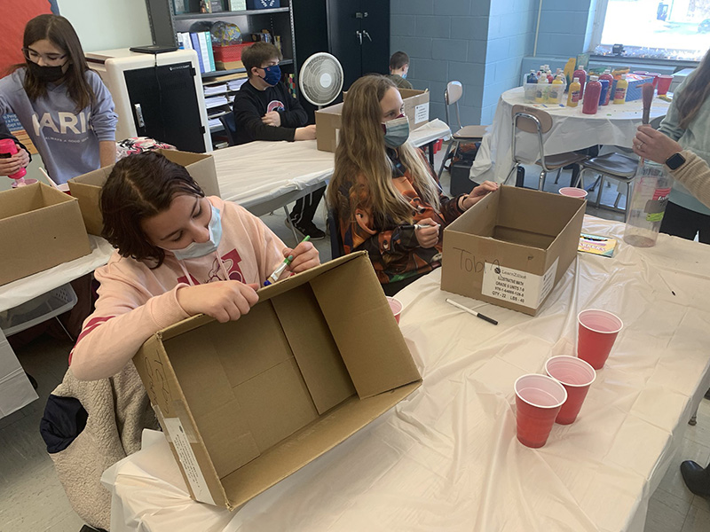 Two middle school students sit at a table with cardboard boxes. They are writing on them There are red cups in front of the boxes with paint in them.