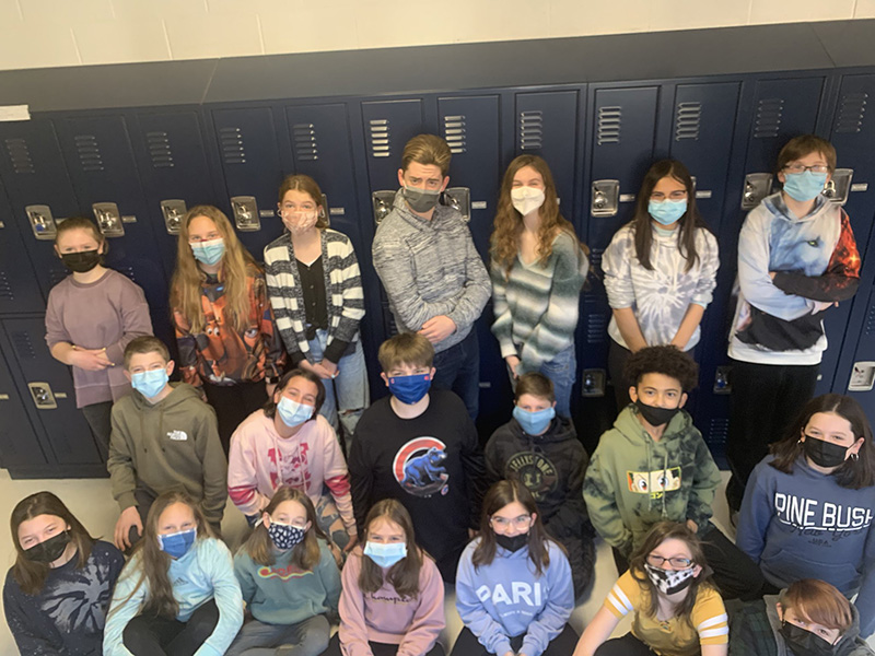 Nineteen middle school students stand in three rows . All are wearing masks. They are standing in front of a row of lockers.