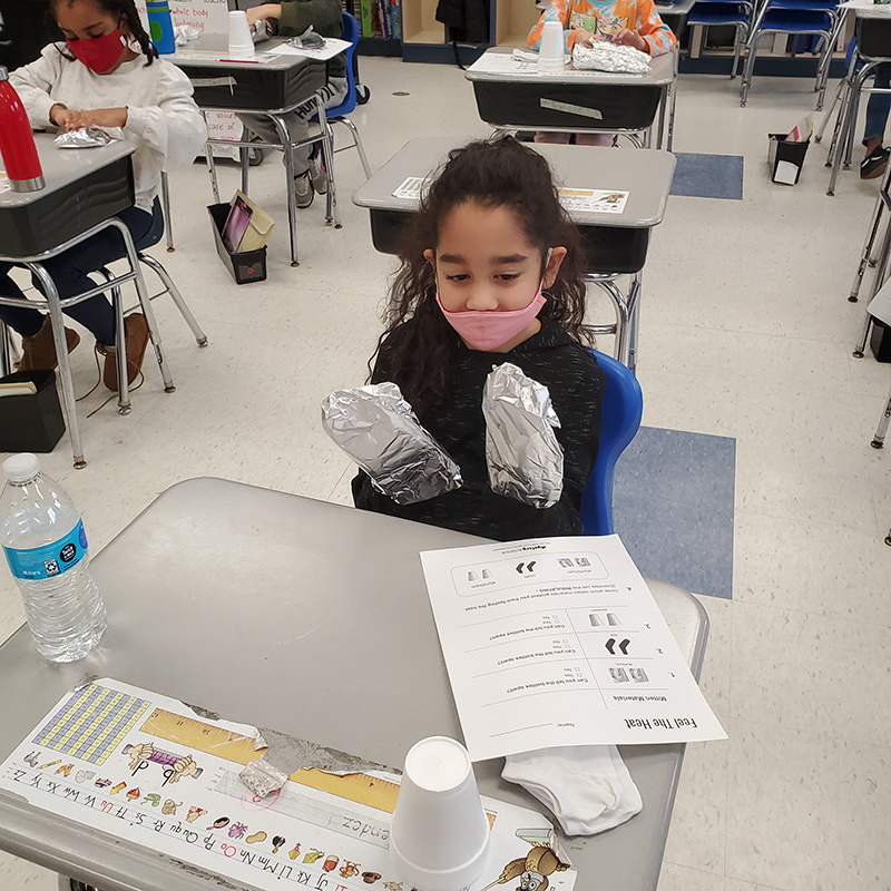 A second-grade girl, with long dark hair wearing a pink mask holds her hands up in front of her. They have aluminum foil on them.