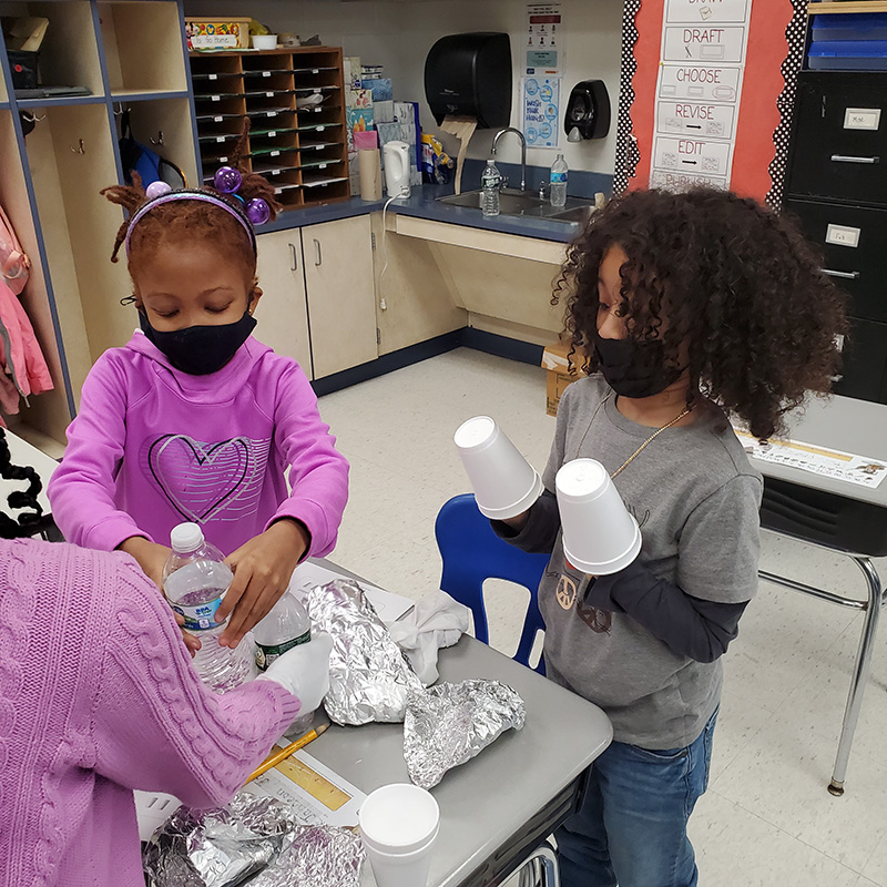 Three second-grade students stand around a desk. One student with shoulder-length dark hair has a styrofoam cup on each hand. Another student in a purple shirt with hairpulled back and wearing a black mask holds a bottle of water.