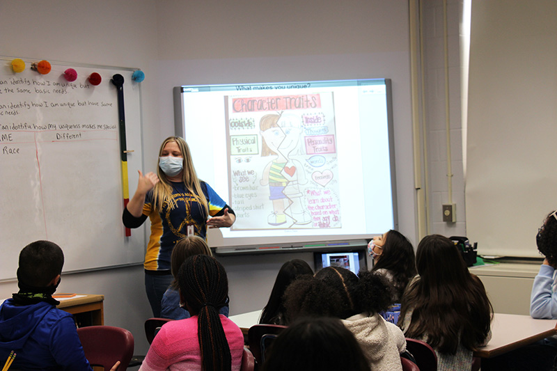 A woman with long blonde hair, wearing a mask and a blue and gold shirt stands in front of a screen in front of a group of fifth grade students.