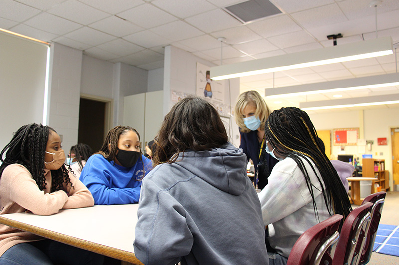 A group of four fifth-grade students sit around a table. A woman with short blonde hair leans over the table talking to them. All are wearing masks.