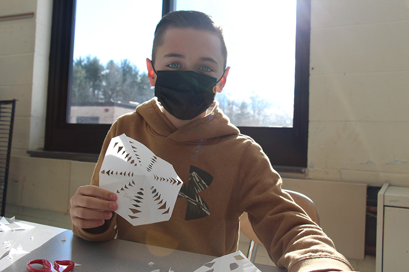 A fourth-grade boy with short dark hair wearing a brown hoodie and black mask holds up a snowflake he cut out of a piece of white paper.