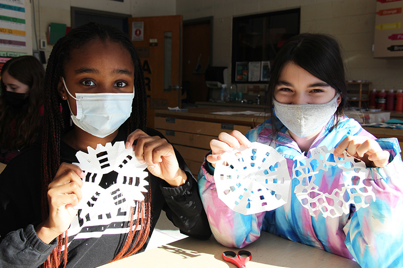 Two fourth-grade girls sit at a table. They are both wearing light blue masks and are holding up paper that is cut into snowflakes. They are smiling behind their masks.