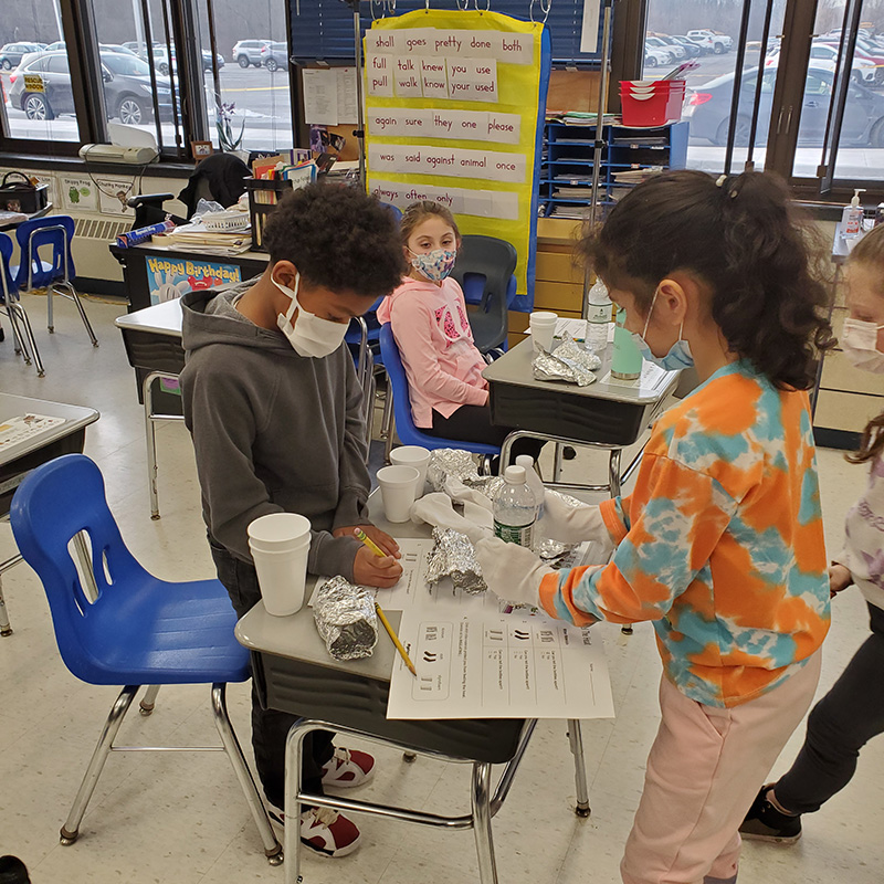 Two second-grade students do a science experiment. The girl on the right has her dark hair pulled back in a ponytail and is wearing an orange and blue shirt and a blue mask. She has white socks on her hands as she picks up a bottle of water. Her classmate, a boy wearing a gray hoodie with short dark hair and wearing a mask, writes on a piece of paper.