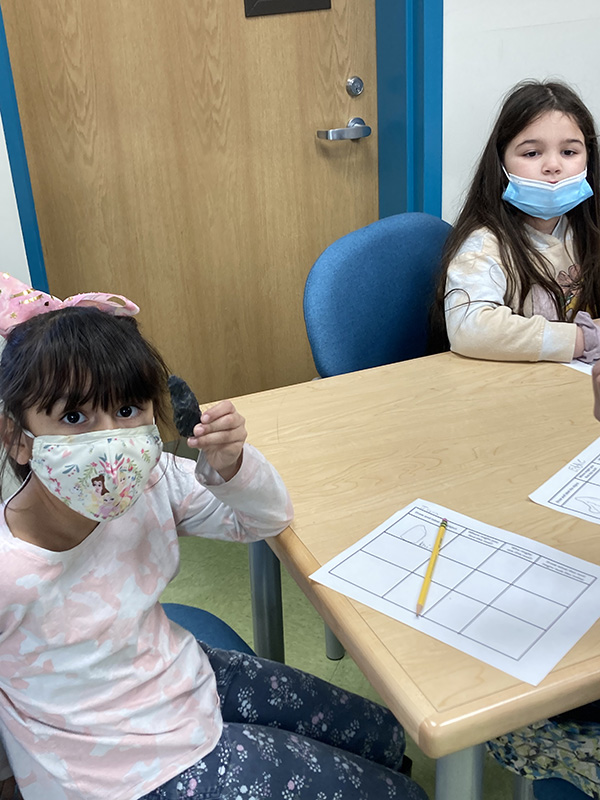 An elementary school-age girl with short dark hair, wearing a pink bow and a patterned face mask, holds up an artifact. On the table is a piece of paper with a graph on it and a pencil on top of it.