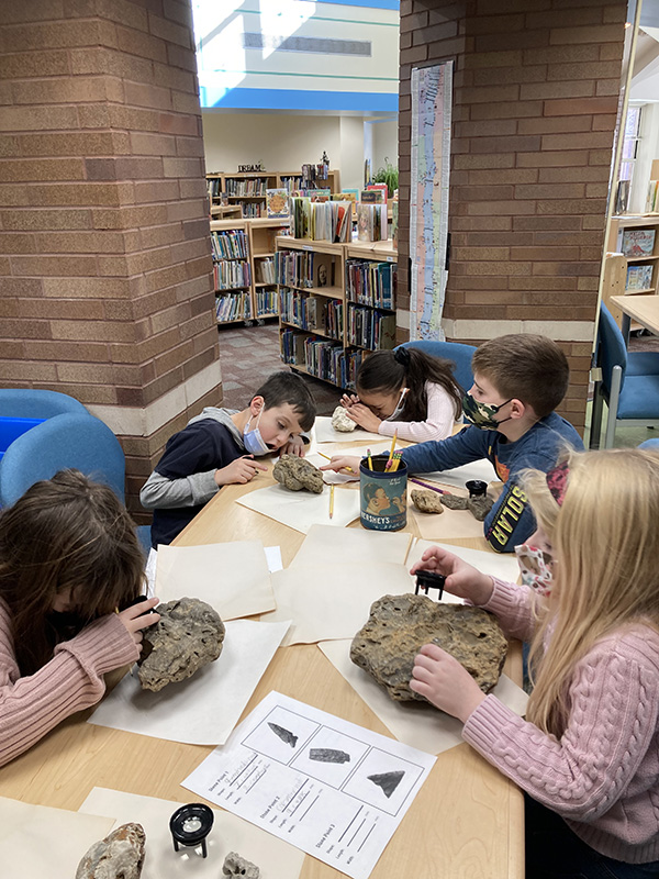 Several elementary-age students sit at a table looking at artifacts. They are also writing down what they are observing.