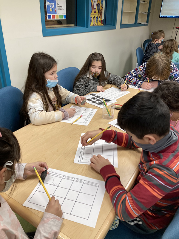 A group of elementary age ids are sitting at a table writing on paper.