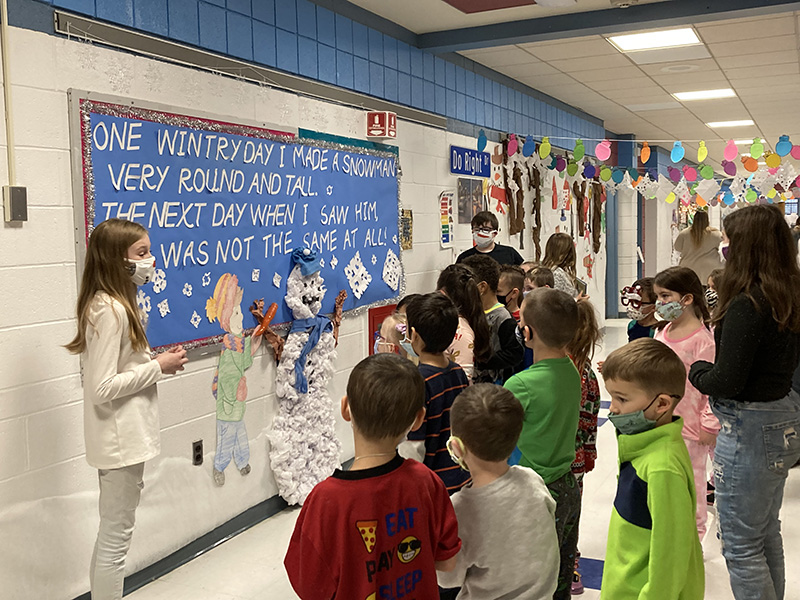 A younger group of elementary students listen as an older student reads a section from a book about snowman. Next to her is a bulletin board in blue with snow scene on it and the words to the story.