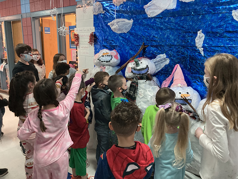 Younger elementary students point to a lifesize scene of snowmen. There is a blue background and the snowmen are made of  large filled plastic bags. All kids have masks on.