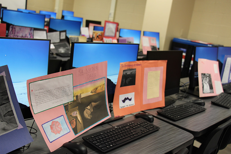 Two posters sit on computer keyboards. The posters contain information about an artist along with a photo of the artist's famous painting and a stamp the students created.