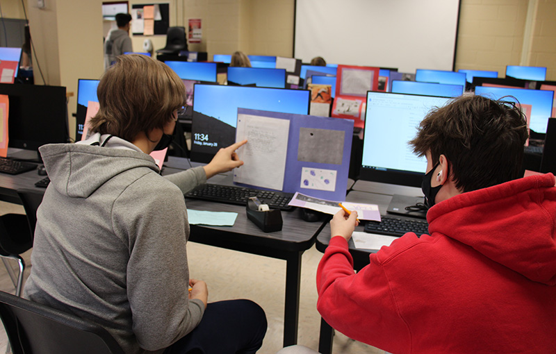 Two high school students work together. One on the eft is pointing at a project he made on construction paper. The other on the right is writing.