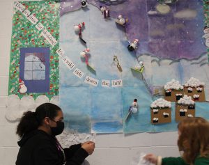 Two older elementary age girls put paper snowflakes onto a bulletin board. The board shows a snow hill with snowmen coming down on sleds. The girls are wearing masks and kneeling to put the snowflakes on the bottom of the hill.