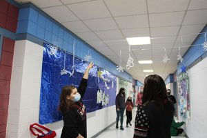 An older elementary-age girl points to the ceiling where paper snowflakes are hanging from string. There are others in the hallway looking up.