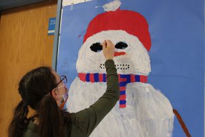 An older elementary-age girl, wearing a gray long-sleeve shirt, a mask and glasses, reaches up to paint the eye of a snowman on blue paper hanging on a wall.