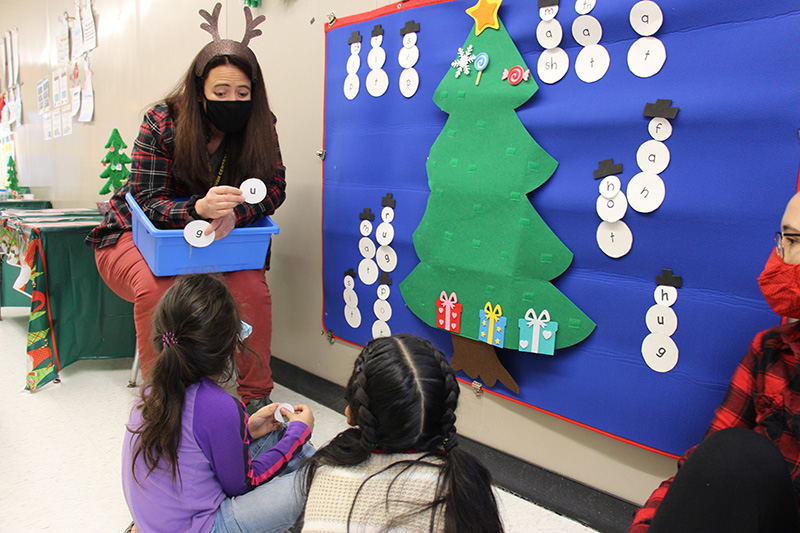 A woman wearing an antler headband and black mask holds a blue bin with papers in it that have letters on them. There is a bulletin board next to her with a blue background and a green Christmas tree on it, surrounded by paper snowmen. Two elementary age girls are in front of the bulletin board looking at the teacher.