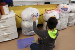 An older elementary-age boy draws a smile on a 3-D snowman made of plastic bags.