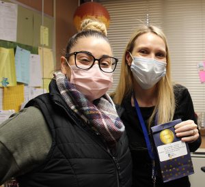 A woman with long blond hair and a mask on holds up a black bag with stars on it. She is standing next to a woman with a bun on top of her head, wearing a black vest, plaid scarf and a pink facemask and glasses.