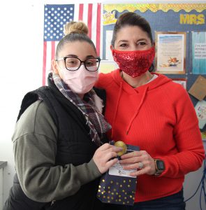 A woman dressed in an orange shirt and orange mask, with dark hair is on the right. She is standing next to a woman with a bun on top of her head, wearing a black vest, plaid scarf and a pink facemask and glasses.